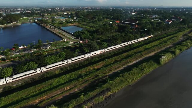 aerial Modern train travels beside green riverside landscape. Train moves along tracks bordered by lush trees and grass. Waterway curves near a calm lake with distant buildings