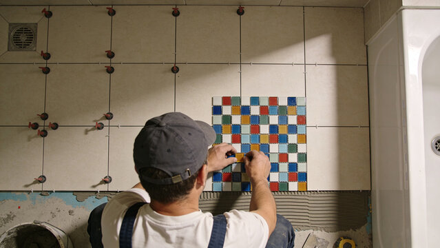 Professional tiler installing colorful mosaic ceramic tiles inside modern bathroom renovation project skilled contractor aligning multicolor square pieces over fresh adhesive mortar detailed craftsman