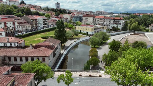 Aerial view of a stone arch bridge and modern curved pedestrian bridge crossing a narrow river channel surrounded by terracotta rooftop buildings and green trees in Portugal