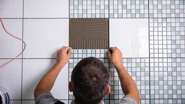 Professional tiler installing ceramic mosaic wall tiles inside modern bathroom renovation project skilled contractor aligning glossy square pieces over fresh adhesive mortar precision craftsmanship co