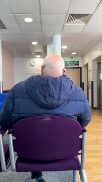 Vertical shot. Man with hearing aid, bone conduction implants sitting on chair in waiting area in hospital, audiology department; rear view, tilt up.