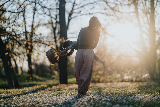A mom spins her child playfully in a sunlit meadow while holding a picnic basket. The joyful scene shows family play and motion in a flowering park at golden hour.