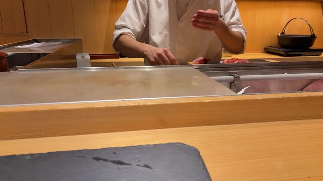 Professional sushi chef preparing fresh tuna fillet at traditional restaurant counter. Hands of Japanese master slicing and seasoning dark red raw fish. 