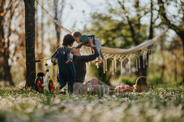 Fototapeta premium Mother plays with her two young children in a sunlit park while holding the baby above her. A relaxed family picnic scene with a hammock, bike and blanket on grassy spring ground.