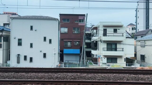 A train passes by typical Japanese residential buildings along the Tokyu Oimachi Line in Setagaya Tokyo. Modern multi-story houses and traditional buildings stand densely packed.