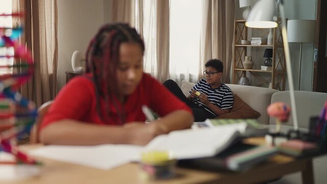 Diligent girl doing homework while her brother plays video games on background