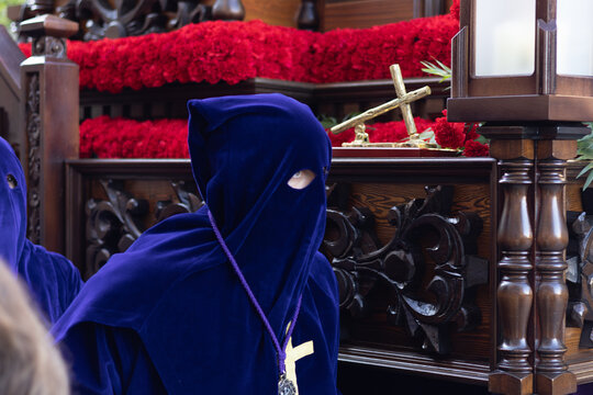 Close-up of a penitent in a blue velvet robe and hood during a Holy Week procession, religious float with red carnations in the background, Asturias.