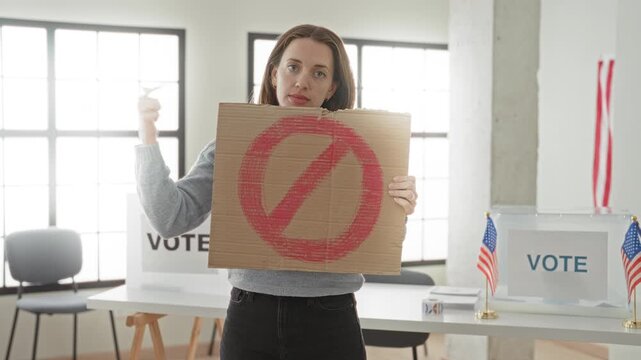 Woman young caucasian holding cardboard sign with red prohibition circle and points finger to sign in a voting building with ballot box and usa flags visible; defiance distrust.