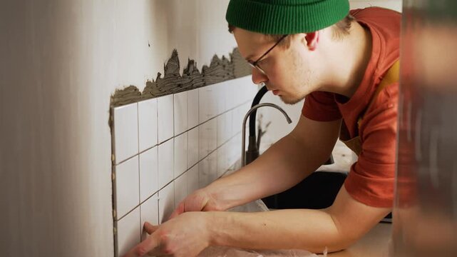 Concentrated male tiler in a green beanie carefully applying small white tiles to fresh adhesive on a wall, installing plastic spacers for even grout gaps during a kitchen remodeling job