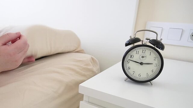 A black alarm clock in his hands by the bed. Smartphone on the bed, the general plan. close-up of the hands setting the alarm clock on the watch and phone.