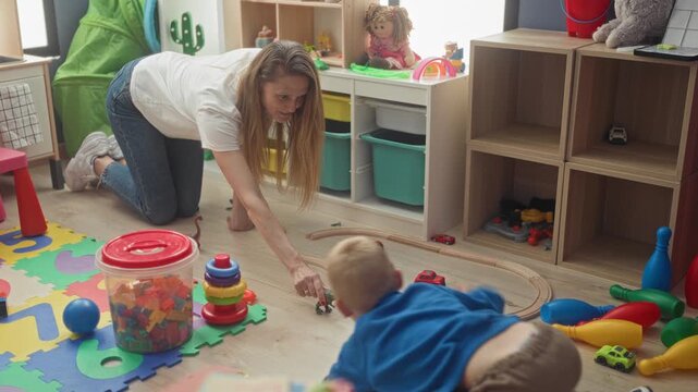 Woman on hands and knees reaching hand with toy dinosaur to boy amid scattered toys in a building play area; nurturing joy.
