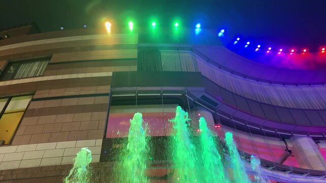 Illuminated fountains jet upwards against a multi-story building facade at night in Fukuoka. Rainbow LED lights align the curved architectural borders of the Canal City Hakata complex.