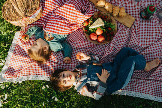 Two young children lie on a red gingham picnic blanket surrounded by fruit and croissants. They relax on the grass during a casual outdoor picnic.