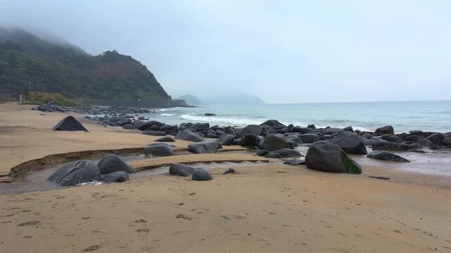 A rocky sandy beach meets the ocean under a cloudy sky in Itoshima Fukuoka. Small waves crash against large black volcanic stones scattered along the coastline. 
