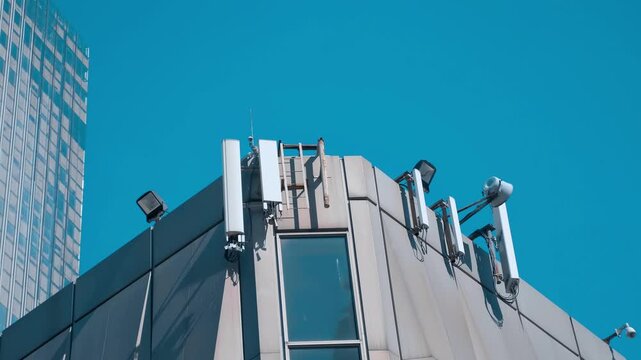 Cellular antennas are positioned high on the corner of a building against a blue sky. Shot in motion