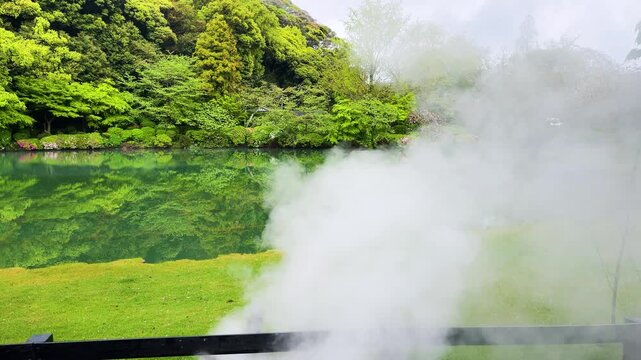 Beautiful Japanese garden pond surrounded by lush green trees and blooming bushes in Beppu. Heavy white steam rises from the thermal hot spring water surface. 