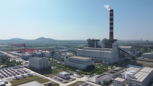 Industrial plant with tall smokestack emitting white plume against clear blue sky and green surroundings