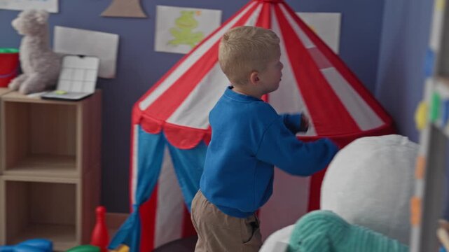 Boy reaching to spin a colorful wall target beside a red and white play tent in a playroom; curiosity exploration.