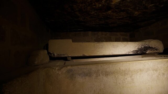 Ancient stone sarcophagus with open lid in dark underground burial chamber inside Mastaba of Ti at Saqqara necropolis, Egypt, dramatically illuminated against rough rock ceiling.