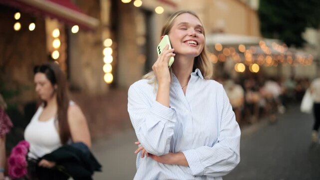 Happy young blonde woman talking on smartphone while standing on a busy city street