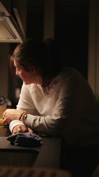 Woman standing at kitchen counter holding head in hands, tired expression, late evening stress and domestic exhaustion moment
