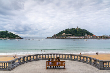 Obraz premium People sitting on a bench enjoying panoramic views of La Concha beach and Monte Urgull in San Sebastian, Spain.