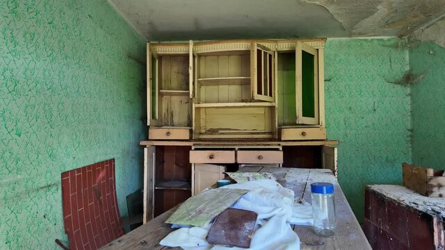 Interior of an abandoned house featuring an old wooden kitchen sideboard with open doors, green walls with peeling wallpaper, cobwebs in corners, wooden table in foreground with a glass jar and rags, 
