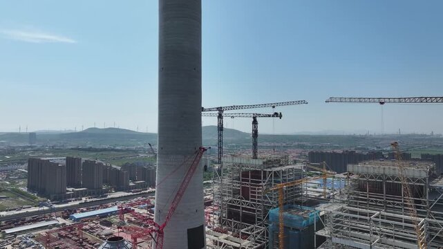 Large industrial chimney and construction site of factory building under development with cranes and steel structures