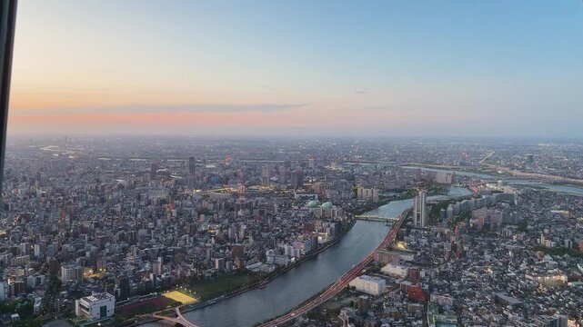 Aerial high-angle footage captures a panoramic evening view of Tokyo city from the Tokyo Skytree observation deck. The camera overlooks the winding Sumida River.
