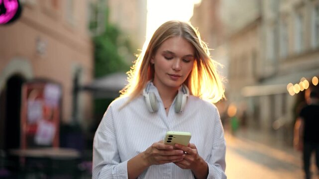 Young blonde woman in headphones texting on smartphone on a city street at sunset