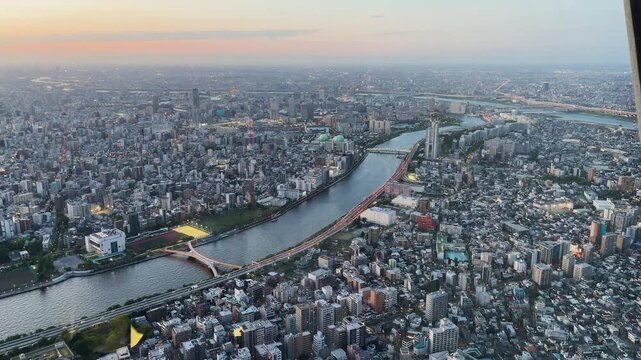 Aerial high-angle footage captures a panoramic evening view of Tokyo city from the Tokyo Skytree observation deck. The camera overlooks the winding Sumida River.