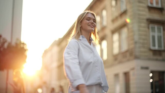 Joyful young blonde woman in pinstripe shirt spinning around with arms outstretched in city at sunset, slow motion