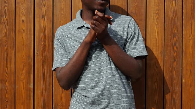 Man stands against wooden wall and claps hands while smiling during a sunny day in the park