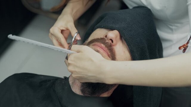 Professional barber trimming beard of male client with scissors and comb during grooming procedure in modern barbershop salon