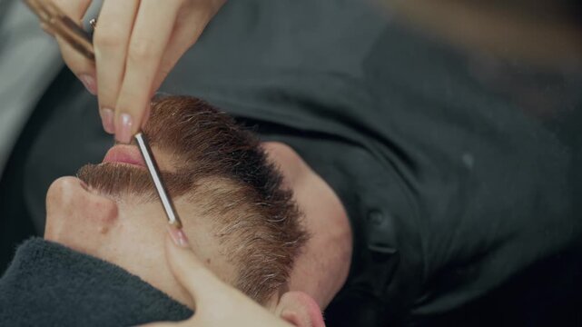 Professional female barber shaving neck of bearded male client with straight razor during grooming procedure in modern barbershop
