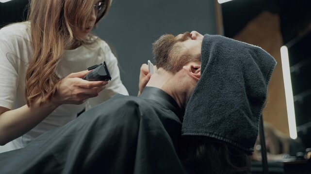 Professional female barber trimming beard of male client with electric clipper during grooming procedure in modern barbershop