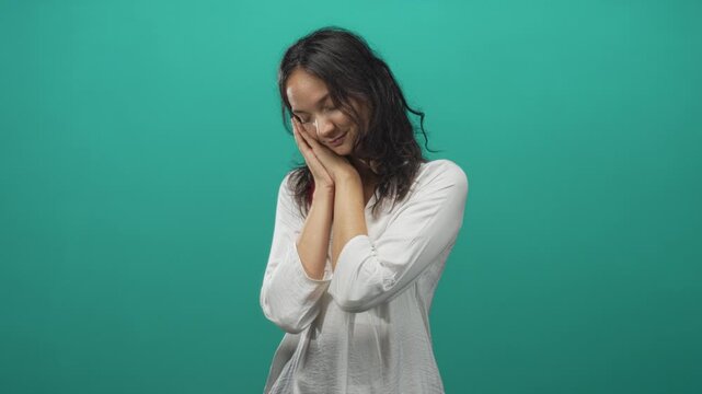 Young hispanic woman resting hands against cheek in a teal studio with white blouse and soft lighting, gentle pose and closed eyes; peaceful rest.