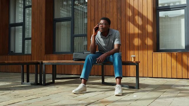Man sitting on bench using phone outside a building in the afternoon sun