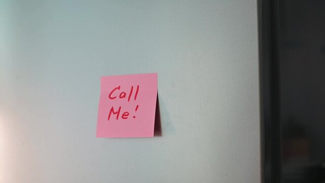 A hand places a pink sticky note with the message Call Me on a smooth surface. This action suggests a reminder or invitation for communication in a home or office environment