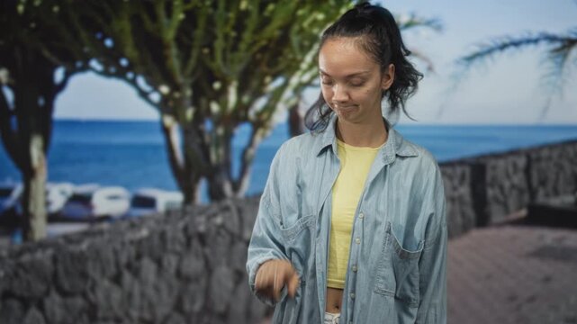 Woman points finger with a slight smirk, wearing denim shirt and yellow tee before an ocean backdrop in studio; mild annoyance.