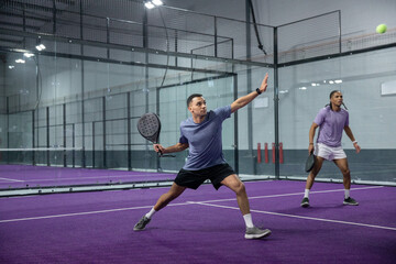 Fototapeta premium Adult men playing padel, foreground player lunging with racket, wearing sportswear on purple court
