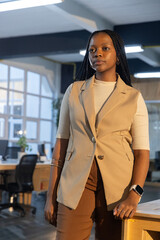 Obraz premium African American woman in 20s leaning on wooden desk in open office, wearing blazer and smartwatch
