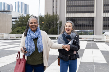 African American senior woman and mature friend walking holding hands on plaza with red tote phone © wavebreak3