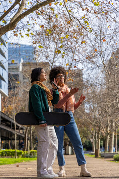 Diverse female friends walking on paved city path holding skateboard and smartphone wearing scarves