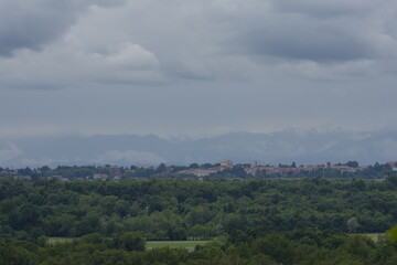 Fototapeta premium A wide landscape shot showing the town of Oleggio visible on the horizon, nestled behind a vast expanse of lush green forest and valley under a bright, cloudy summer sky.