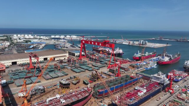 Large shipyard port with ongoing ship construction and heavy lifting operations under clear blue sky