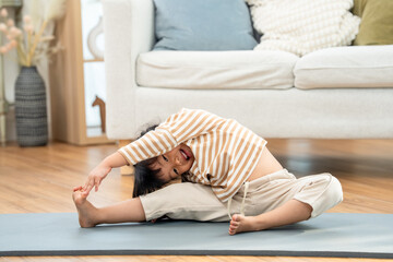 Obraz premium Asian preschool girl bends sideways on yoga mat, reaches toward foot with playful smile, practicing flexibility, balance, body control, stretching exercise, child wellness routine at home indoors.