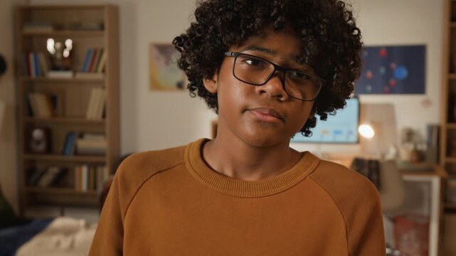 Medium close-up portrait of teenage curly-haired African American boy in geeky glasses blowing dust off textbook to prepare to go back to school and start new semester after summer vacation