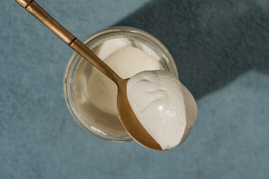 Top view of thick white yogurt or cream cheese or sour cream in glass jar with a golden spoon on a blue background. Minimalist healthy breakfast concept with natural sunlight and shadows