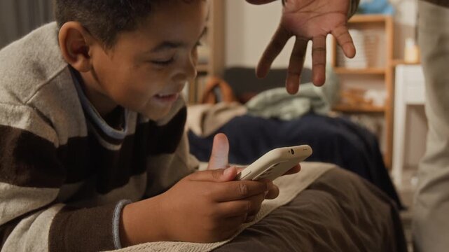 Medium close-up of young African American boy browsing or playing on smartphone in bedroom, arguing and pleading with father removing his smartphone, for parent control over screen time and wellbeing